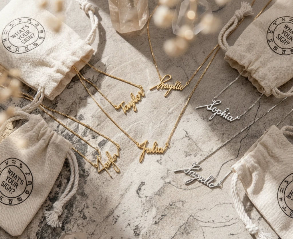 A styled flat-lay of personalized name necklaces and a matching name ring, with a hand visible wearing the ring. The jewelry is arranged on a textured stone surface with cream drawstring pouches, crystals, and dried flowers.